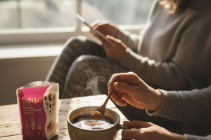 A close-up shot of a person adding a spoonful of MISO powder into a steaming hot drink in a mug. In the foreground, an open pink and black pouch of MISO powder is visible. In the slightly blurred background, another person sits next to the mug holder, looking at a tablet or phone near a bright window.