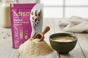A close-up of a pink and white pouch of MISO powder superfood displayed on a wooden table next to a pile of the light-colored powder. A small wooden scoop rests in the powder pile, and a dark green bowl of prepared MISO broth or soup is shown on the right, highlighting the product's form and application.