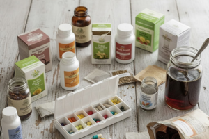 A collection of various bottles, boxes, and jars of pills, capsules, teas, and liquids for digestive relief (including a full pill organizer and tea bags) scattered on a wooden table, representing the numerous and often unsuccessful supplement-based remedies tried for chronic constipation.