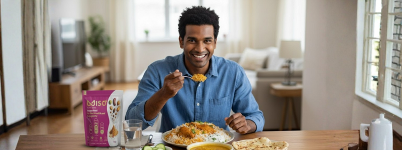 A smiling man sits at a dining table in a bright, modern living space, enjoying a meal of rice (likely biryani or pulao), dal, and naan. He is holding a spoonful of food towards the camera. On the table, to his left, is a prominent pink and white pouch of MISO powder, suggesting its easy integration into daily Indian meals.
